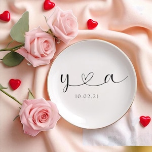 May include: A white plate with a gold rim features the letters "y & a" with a heart symbol, and the date "10.02.21" in black script. The plate is surrounded by pink roses and red heart-shaped candies on a pink fabric.