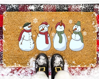 May include: A rectangular doormat with four cartoon snowmen, each wearing a hat and scarf. The mat has a textured, tan surface with a red and black plaid border. Black shoes with gold buckles are visible at the bottom.
