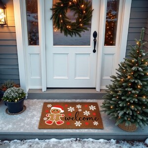 May include: A festive doormat featuring a gingerbread man in a Santa hat, with the word "welcome" and snowflakes. A small Christmas tree and a wreath are also visible, creating a welcoming holiday scene.