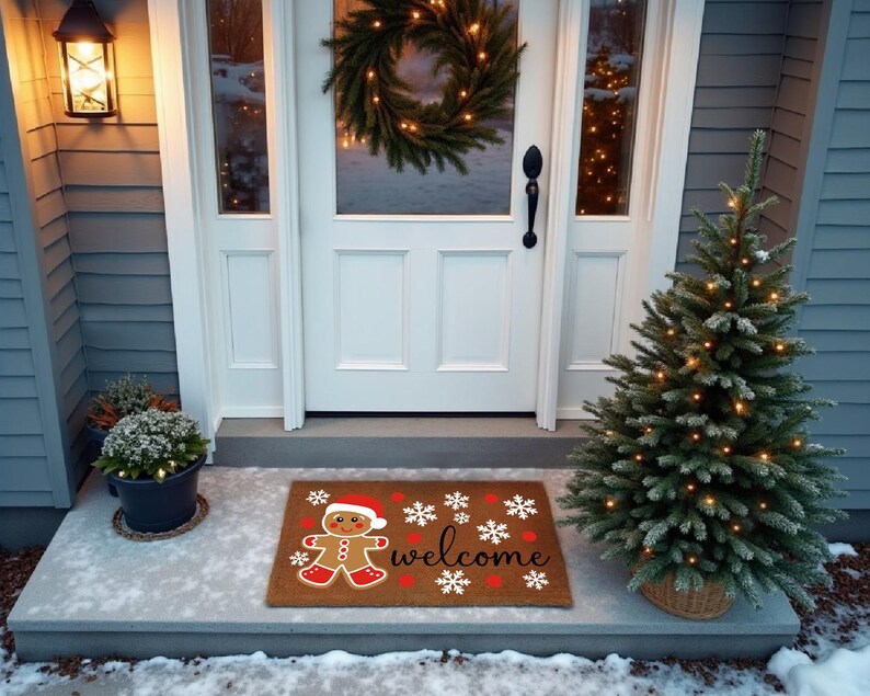 May include: A festive doormat featuring a gingerbread man in a Santa hat, with the word "welcome" and snowflakes. A small Christmas tree and a wreath are also visible, creating a welcoming holiday scene.