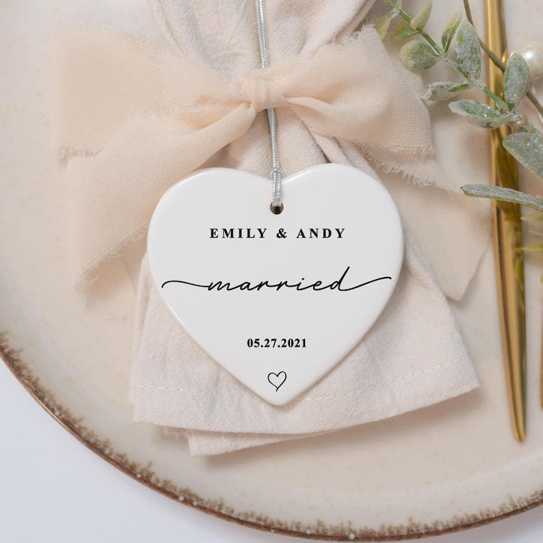 K&ouml;nnte beinhalten: Ein wei&szlig;es herzf&ouml;rmiges Ornament mit den Namen "EMILY & ANDY" und dem Wort "married" in Schreibschrift. Das Datum "05.27.2021" und ein Herzsymbol sind ebenfalls vorhanden. Das Ornament h&auml;ngt an einem silbernen Faden.