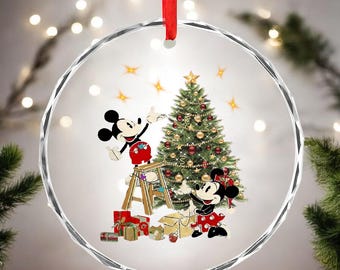 May include: A clear, round Christmas ornament with a red ribbon. The ornament features Mickey and Minnie Mouse decorating a Christmas tree. Mickey is on a ladder, and Minnie is on the ground. Presents are at the base of the tree.