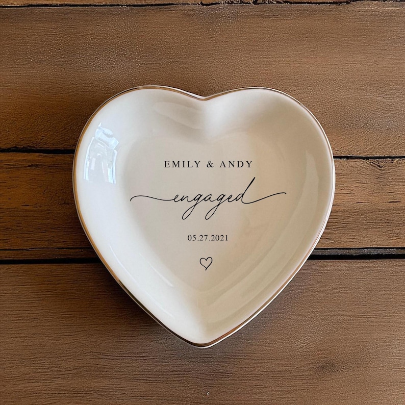 May include: A heart-shaped, white ceramic trinket dish with a gold rim. The dish is inscribed with "EMILY & ANDY" above the word "engaged" in cursive, and the date "05.27.2021" with a heart symbol below.