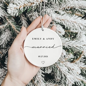 K&ouml;nnte beinhalten: Runder, wei&szlig;er Keramik-Weihnachtsschmuck mit den Namen "EMILY & ANDY" und dem Wort "married" in Schreibschrift. Das Datum "05.27.2021" und ein Herzsymbol sind auf dem Ornament aufgedruckt. Eine silberne Schnur ist zum Aufh&auml;ngen befestigt.
