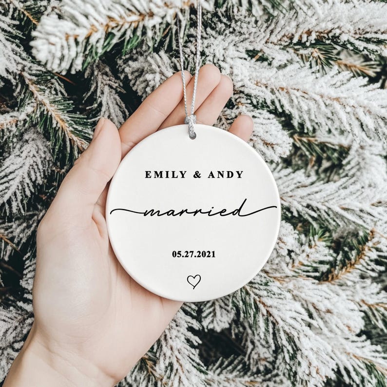 K&ouml;nnte beinhalten: Runder, wei&szlig;er Keramik-Weihnachtsschmuck mit den Namen "EMILY & ANDY" und dem Wort "married" in Schreibschrift. Das Datum "05.27.2021" und ein Herzsymbol sind auf dem Ornament aufgedruckt. Eine silberne Schnur ist zum Aufh&auml;ngen befestigt.