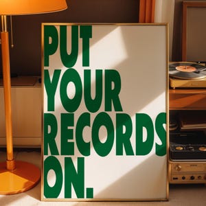 May include: A framed poster with the words "PUT YOUR RECORDS ON." in large, green, rounded letters. The poster has a white background and a gold frame. The image is in a room with a record player and a floor lamp.