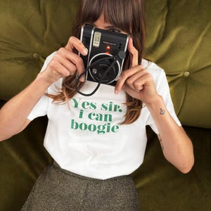 May include: A black Instax Mini 40 instant camera with a black strap is held up. The person is wearing a white t-shirt with the green text "Yes sir, I can boogie". An anchor tattoo is visible on the arm.
