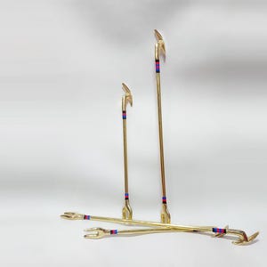 Peut inclure: Quatre outils en métal doré avec un motif en forme d'oiseau en haut et un motif en forme de main en bas. Chaque outil a une rayure rouge et bleue près du sommet. Les outils sont disposés sur une surface blanche.