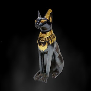 May include: A black cat statue with gold accents, seated and facing left. The statue features a detailed gold collar and headpiece, with intricate designs. The cat's eyes are also highlighted in gold, set against a black background.