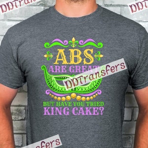 Peut inclure: Un t-shirt gris avec un motif vert, violet et or qui dit "ABS ARE GREAT BUT HAVE YOU TRIED KING CAKE?"
