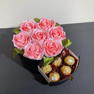 May include: A black gift box with a drawer filled with gold wrapped chocolates. The top of the box is decorated with a bouquet of pink satin roses.
