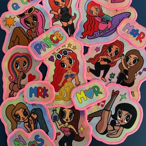 May include: A collection of colorful iron-on patches featuring cartoon girls with different hairstyles and outfits. The patches have pink stitching around the edges and include text like "Q Chimba", "Parce", "Mrk", "Mor", "Cur Del", and "Adios Mi".