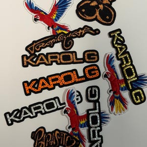 May include: A collection of embroidered patches featuring a red, blue, and yellow parrot, the name "KAROL G" in various fonts, and the word "PAPASITO". The patches have glitter accents.