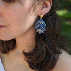 May include: A close-up of a person wearing a pair of leaf-shaped earrings. The earrings are blue with gold accents and have a layered design. The earrings are attached to a small gold stud.