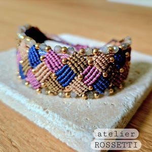 May include: A multi-coloured braided bracelet with gold beads and iridescent stones. The bracelet is made with a chevron pattern and has a light brown, pink, and blue colour scheme. The text "atelier ROSSETTI" is visible in the image.