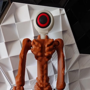 Puede incluir: Una figura articulada marrón con un solo ojo blanco con un anillo rojo y una pupila negra. La figura tiene múltiples miembros articulados y está posada con los brazos extendidos.