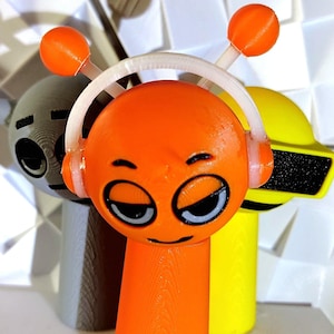May include: Three 3D printed figurines in orange, yellow and grey with cartoon faces. The orange figurine has a white headband and two antennae. The yellow figurine has a black visor. The grey figurine has a black eye.