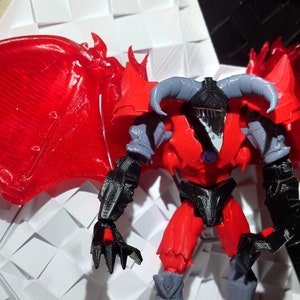 May include: A red and black action figure with wings, horns, and a purple gem on its chest. The figure is standing on a white surface.