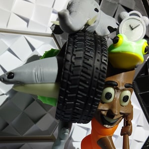 May include: A whimsical sculpture featuring a shark, elephant, frog, and cartoon character with a tire, clock, and blue sneaker. The sculpture is made of various materials and has a playful, surreal design. The color palette includes gray, black, orange, green, and blue.