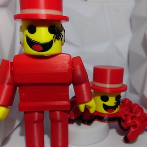 Könnte beinhalten: Eine rote, bewegliche Figur mit gelbem Kopf, schwarzen Augen und einem lächelnden Mund, die einen roten Zylinder trägt. Eine kleine schwarze Spinne ist am Gesicht der Figur befestigt. Eine weitere ähnliche Figur befindet sich im Hintergrund.