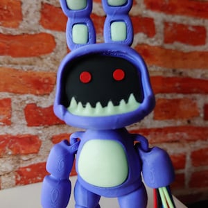 May include: A purple figurine of a rabbit-like character with a black face, red eyes, and a white-toothed grin. The figure has large ears with light green accents and exposed wires on one arm. The character has a light green oval on its chest.