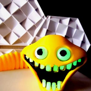 Puede incluir: Una escultura amarilla y caricaturesca con una amplia sonrisa dentuda y grandes ojos verdes. La boca y los dientes brillan en verde. Una estructura geométrica blanca está en el fondo.