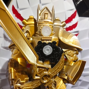 May include: A gold-coloured, 3D printed action figure of a knight wearing armour and holding a sword. The knight has a black circular object on its chest.