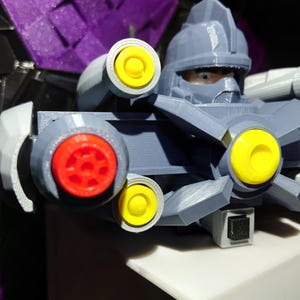 May include: A gray plastic toy robot with a yellow and red face and multiple yellow and gray guns. The robot is wearing a gray helmet with a black visor.