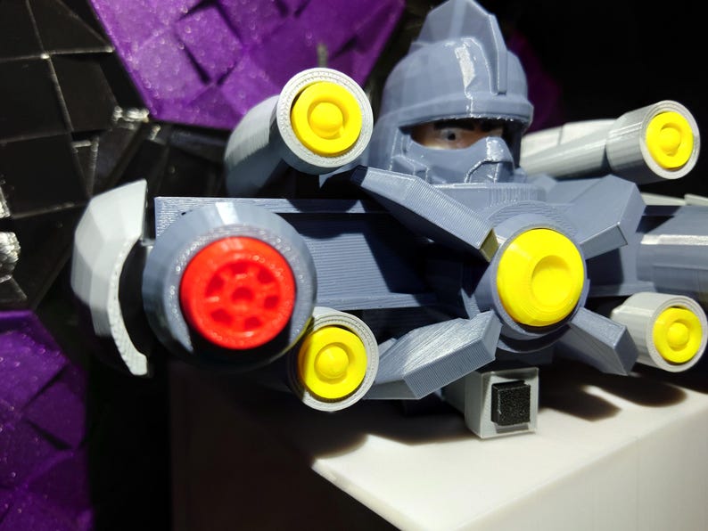 May include: A gray plastic toy robot with a yellow and red face and multiple yellow and gray guns. The robot is wearing a gray helmet with a black visor.