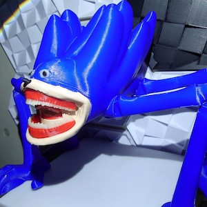 May include: A blue, stylized creature with a large open mouth revealing teeth. The figure has long limbs and a spiky head. The creature is posed on a white surface, with a geometric background of black and white shapes.
