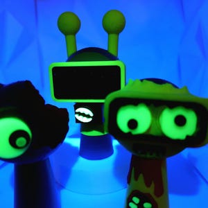 May include: Three glow-in-the-dark figurines, one green robot with a square head and two antennas, one green zombie with two eyes and a mouth, and one black and green creature with one eye.