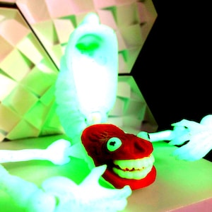 May include: A neon-toned, abstract sculpture featuring a red, smiling face with white teeth and green eyes. The face is attached to a white skeletal structure. The background includes geometric shapes and a black area.