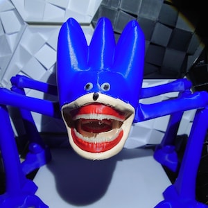 May include: A blue, abstract sculpture with a cartoonish face and large open mouth. The sculpture has four legs and three tall, pointed protrusions on top of its head. The mouth features red lips and white teeth. The background is a geometric pattern.
