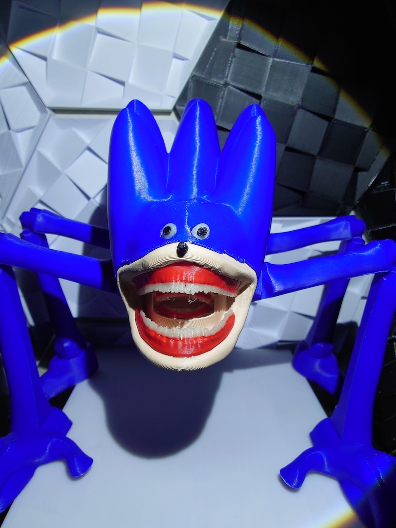 May include: A blue, abstract sculpture with a cartoonish face and large open mouth. The sculpture has four legs and three tall, pointed protrusions on top of its head. The mouth features red lips and white teeth. The background is a geometric pattern.