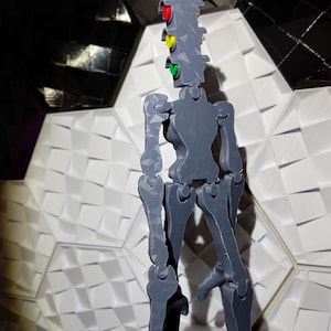 May include: A gray articulated figure with a traffic light head. The traffic light has red, yellow, and green lights. The figure is standing on a white textured surface.