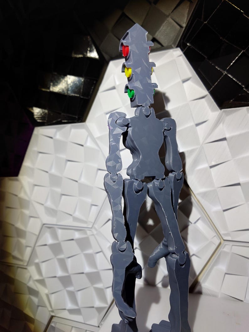 May include: A gray articulated figure with a traffic light head. The traffic light has red, yellow, and green lights. The figure is standing on a white textured surface.