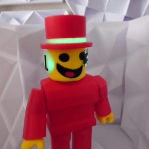 Op de afbeelding: Een rode figuur met een gele kop en handen, met een rode hoge hoed met een groene band. De figuur heeft een lachend gezicht en staat voor een witte geometrische achtergrond. De armen en benen van de figuur zijn recht en blokkerig.