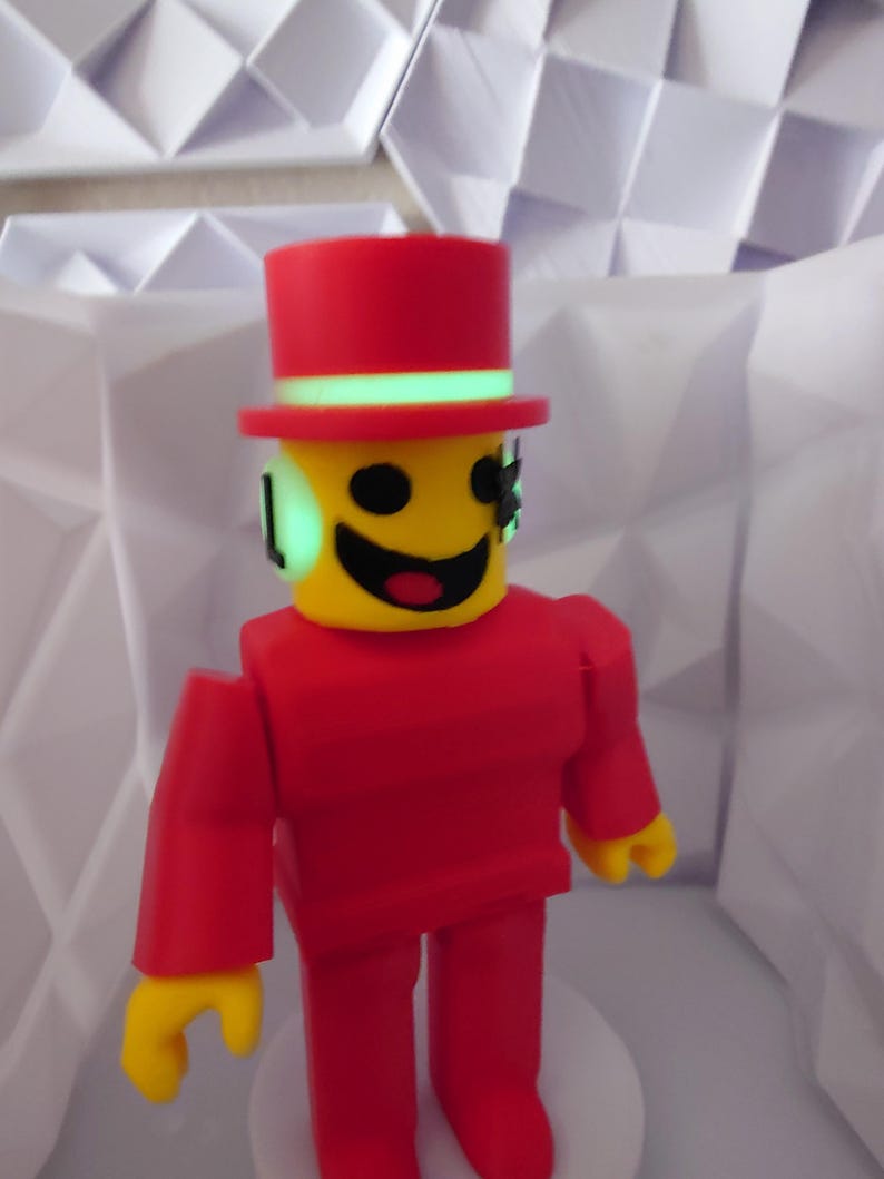 Op de afbeelding: Een rode figuur met een gele kop en handen, met een rode hoge hoed met een groene band. De figuur heeft een lachend gezicht en staat voor een witte geometrische achtergrond. De armen en benen van de figuur zijn recht en blokkerig.