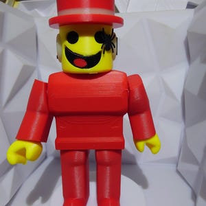 Op de afbeelding: Een rood en geel 3D-geprint figuur met een hoge hoed en een spin op het gezicht. Het figuur heeft een gele kop met een lachend gezicht en een rode romp, armen en benen. Het staat op een witte basis.