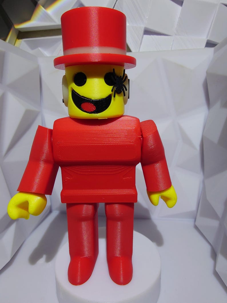 Op de afbeelding: Een rood en geel 3D-geprint figuur met een hoge hoed en een spin op het gezicht. Het figuur heeft een gele kop met een lachend gezicht en een rode romp, armen en benen. Het staat op een witte basis.
