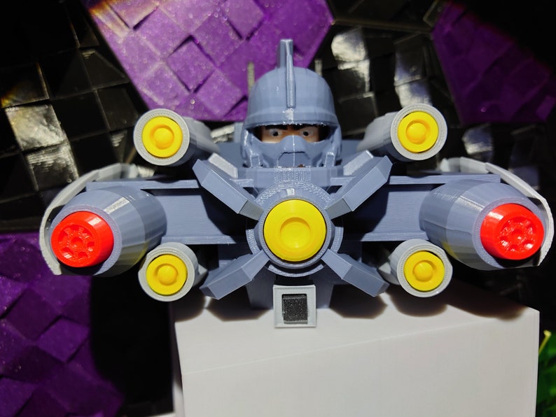 May include: A gray and white robot toy with a helmet and multiple yellow and red cannons. The robot is posed with the cannons facing forward.