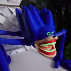 May include: A blue, stylized creature with a large, open mouth revealing glowing green teeth and red gums. The creature has large, green eyes and long, blue limbs. The figure is posed on a white surface, with a textured background of white and black geometric shapes.