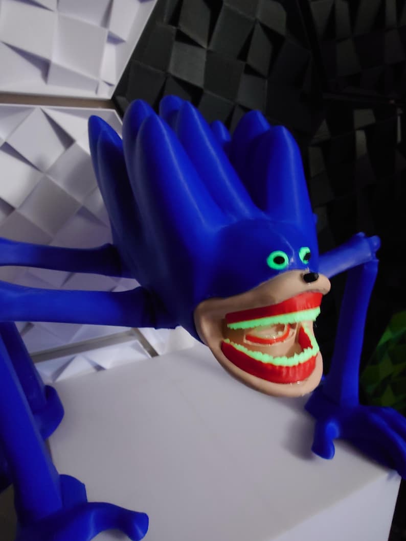May include: A blue, stylized creature with a large, open mouth revealing glowing green teeth and red gums. The creature has large, green eyes and long, blue limbs. The figure is posed on a white surface, with a textured background of white and black geometric shapes.