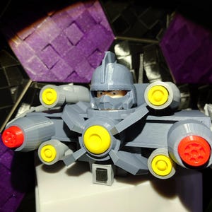 May include: A gray 3D printed robot toy with yellow and red details. The robot has a helmet with a visor and multiple cannons.
