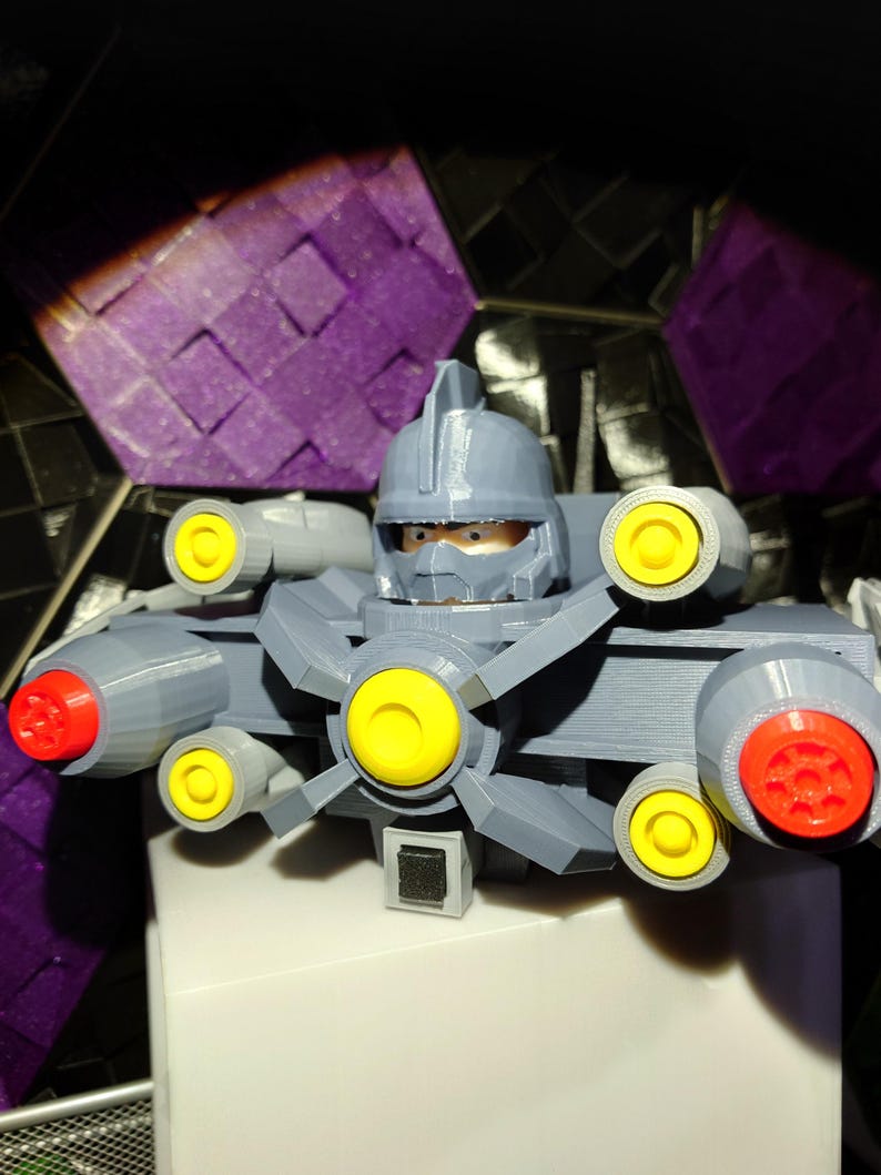 May include: A gray 3D printed robot toy with yellow and red details. The robot has a helmet with a visor and multiple cannons.