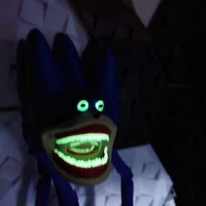 May include: A blue, stylized creature with glowing green eyes and a wide, open mouth revealing glowing teeth. The creature has long, thin blue legs and a dark background.
