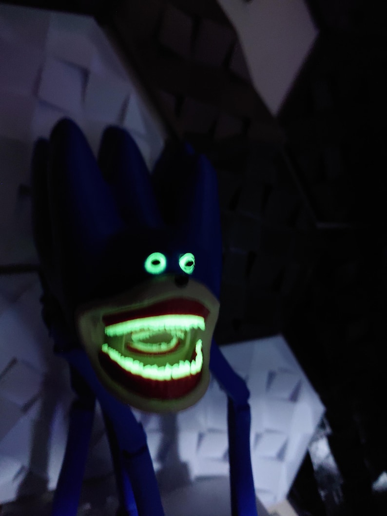 May include: A blue, stylized creature with glowing green eyes and a wide, open mouth revealing glowing teeth. The creature has long, thin blue legs and a dark background.