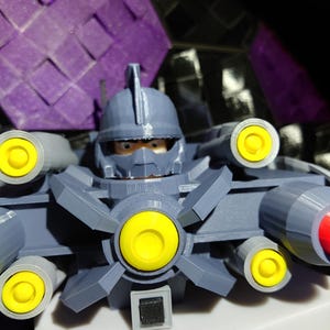 May include: A gray 3D printed toy robot with a yellow helmet and multiple yellow and red cannons. The robot has a black visor and a black square on its chest.