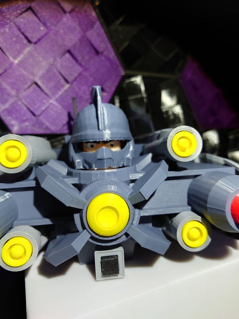 May include: A gray 3D printed toy robot with a yellow helmet and multiple yellow and red cannons. The robot has a black visor and a black square on its chest.