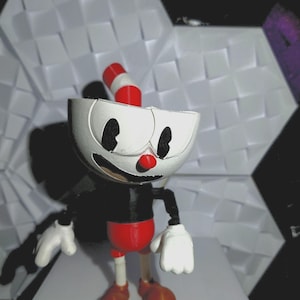 以下が含まれることがあります： 漫画のカップキャラクターの3Dプリントフィギュア。カップは白いボディ、黒い目、赤い鼻をしています。赤いショートパンツ、黒い胴体、茶色の靴を履いています。カップのストローは赤と白のストライプです。